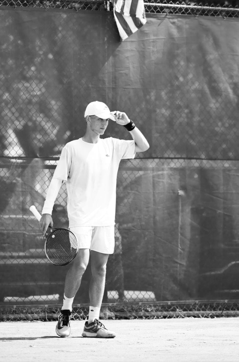 Graeme Angus, founder of Acemore, on a tennis court, touching the brim of his cap, in black and white